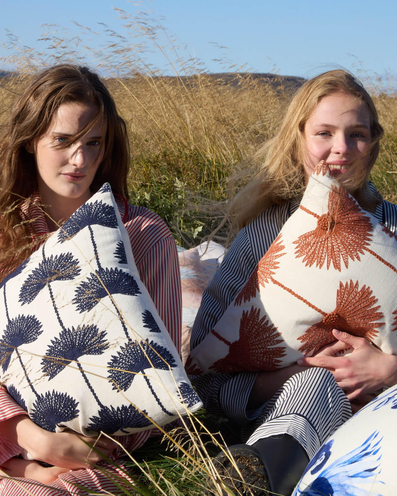 Two women sitting in sunny golden grass field holding palm leaf embroidered throw pillows navy off white and sienna off white variants  with blue sky and rolling hills background