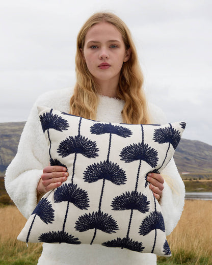 Woman holding palm leaf embroidered throw pillow navy off white variant outdoors wearing white sherpa sweater with golden grass field and overcast highland landscape in background