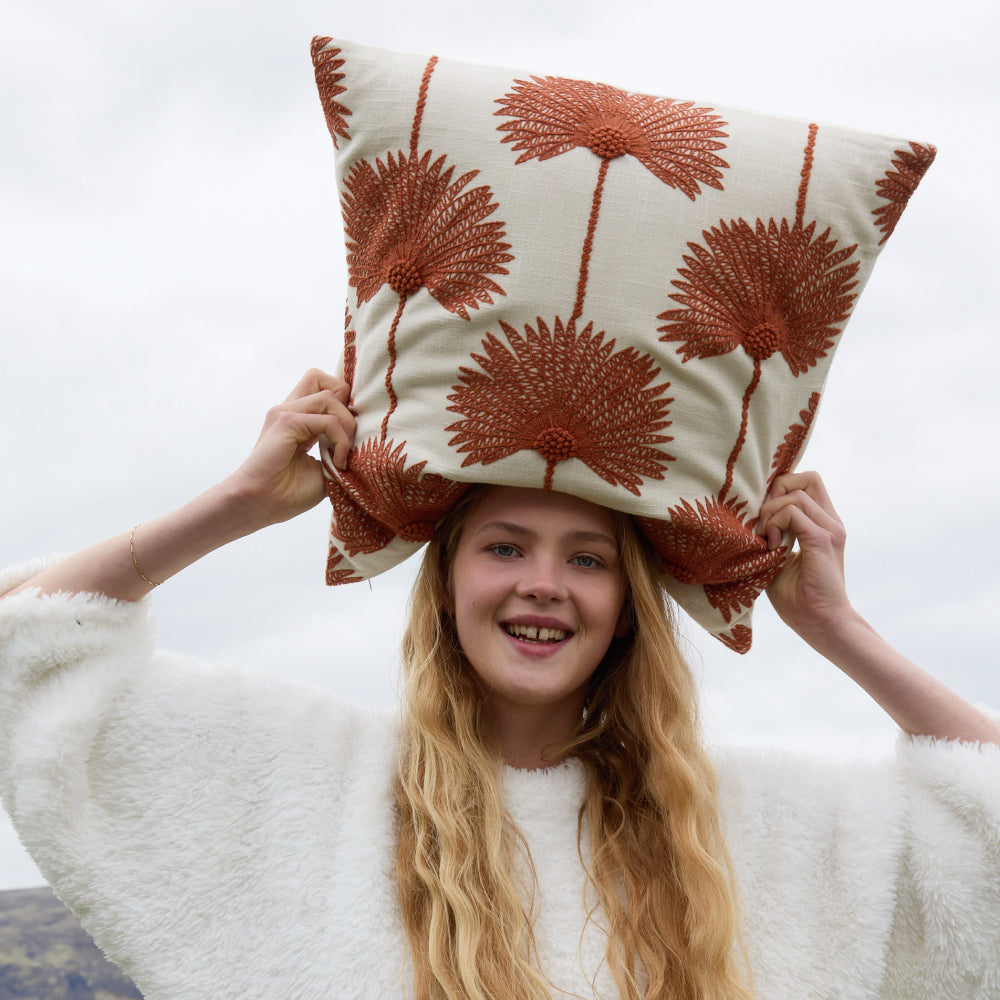 Smiling woman balancing palm leaf embroidered throw pillow sienna off white variant on her head outdoors wearing white sherpa wrap with overcast sky background