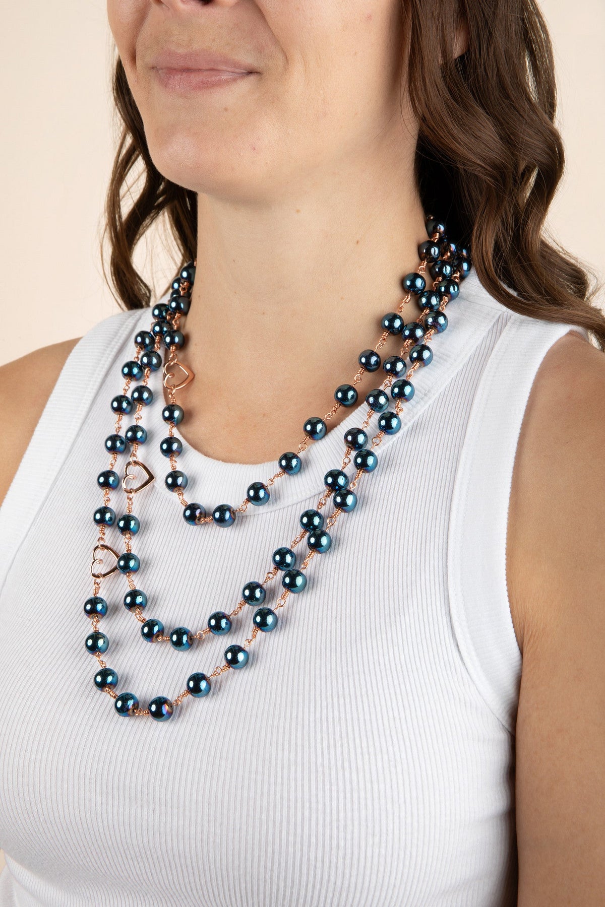 Model wearing the Celeste blue hematite layered necklace over a white ribbed sleeveless top, showing three beaded strands and open heart accents.