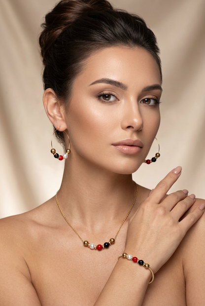 Model wearing the His-Story Majorca pearl bar necklace with coordinating hoop earrings and a bangle bracelet featuring gold, white, red, and black beads.