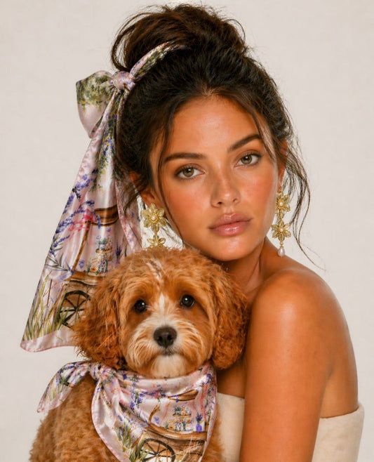 Model wearing the Lavender Bloom silk scarf tied around a high bun, holding a dog with a matching blush garden-cart scarf around its neck.