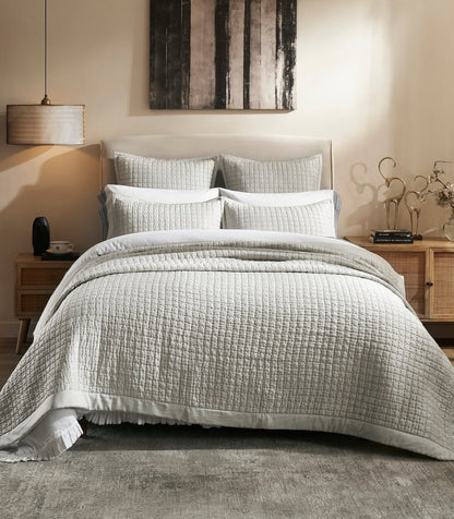 Malibu Mist quilted silk coverlet in a pale gray tone, showing square stitching and a smooth border on a bed between wooden nightstands