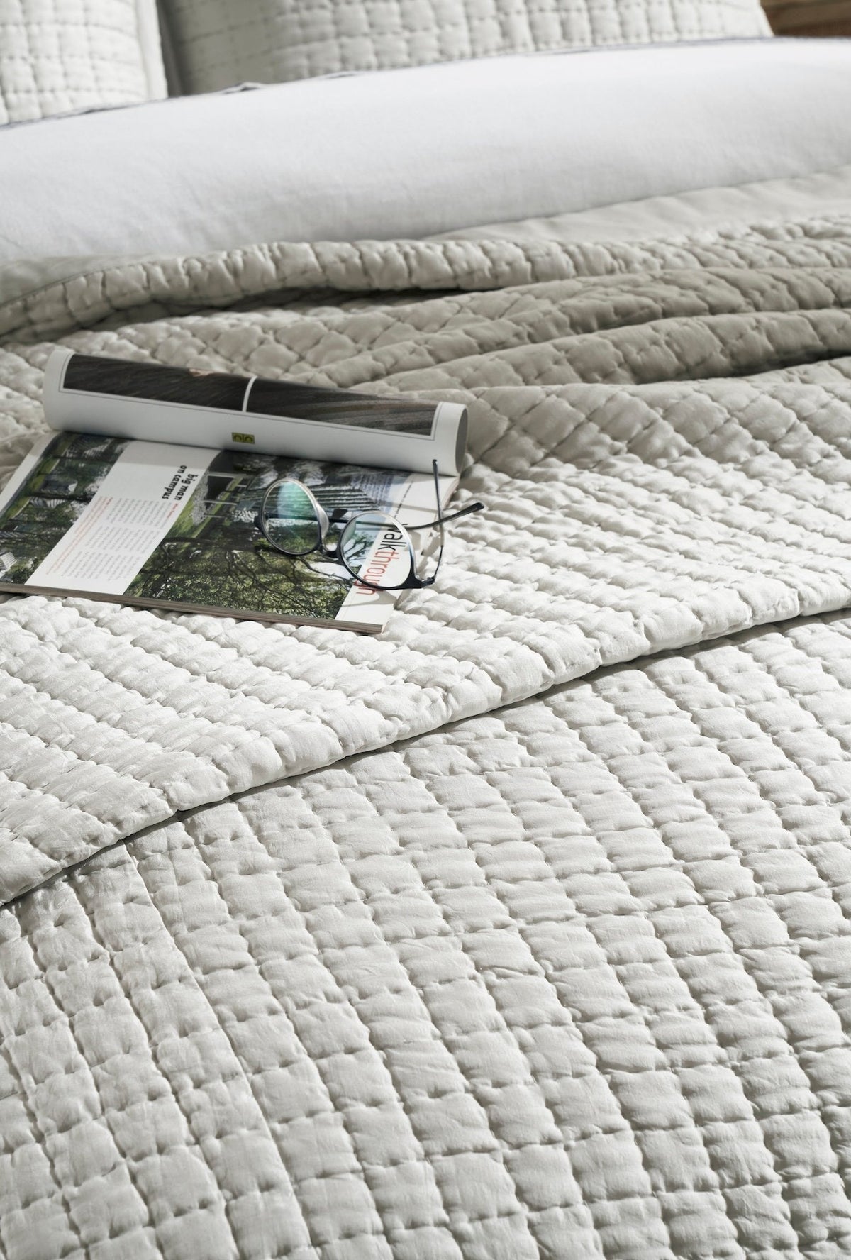 Close-up of the Malibu Mist silk coverlet showing pale gray square quilting and a soft sheen, with an open book and eyeglasses resting on top.