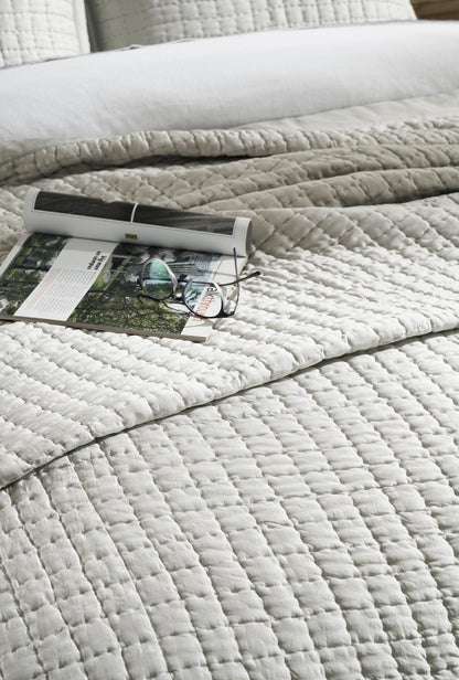 Close-up of the Malibu Mist silk coverlet showing pale gray square quilting and a soft sheen, with an open book and eyeglasses resting on top.