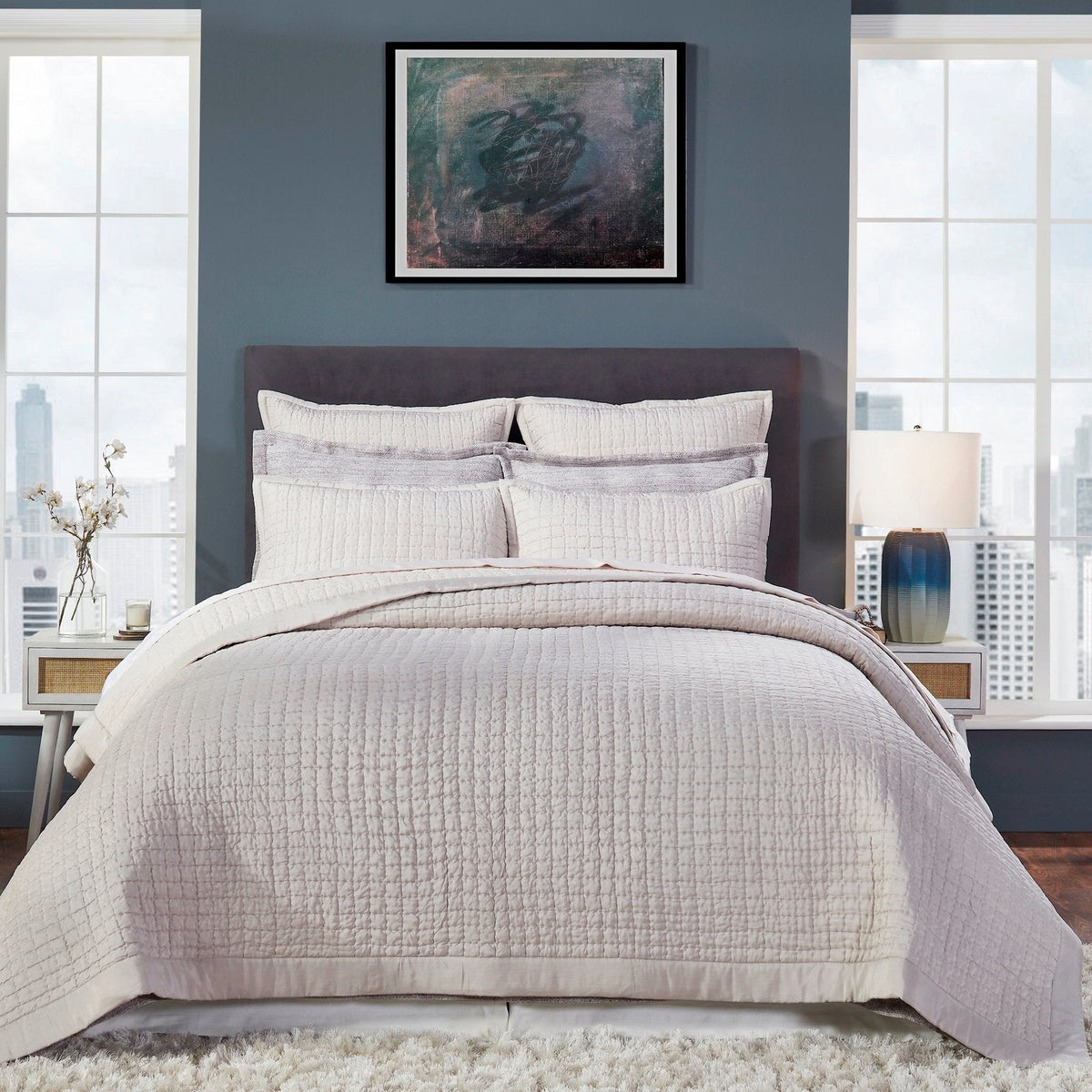 Malibu Oyster quilted silk coverlet in a pale warm beige tone, showing square stitching and a smooth border on a bed against a blue-gray wall.⠋