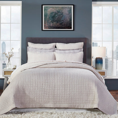 Malibu Oyster quilted silk coverlet in a pale warm beige tone, showing square stitching and a smooth border on a bed against a blue-gray wall.⠋