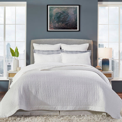 Malibu Pearl quilted silk coverlet spread across a bed, showing its soft ivory tone, square stitching, and smooth border in a gray-walled bedroom.