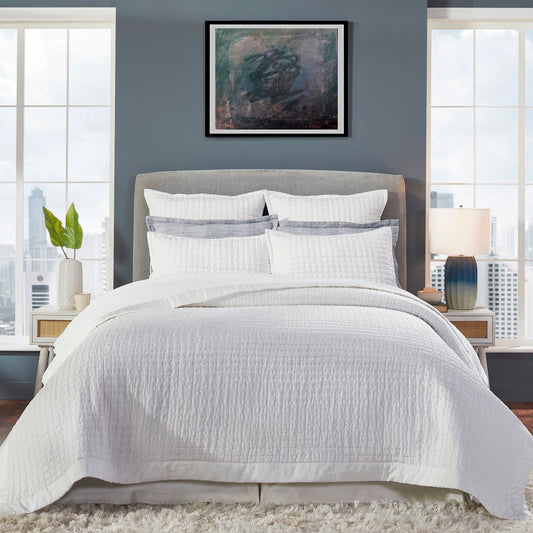 Malibu Pearl quilted silk coverlet spread across a bed, showing its soft ivory tone, square stitching, and smooth border in a gray-walled bedroom.