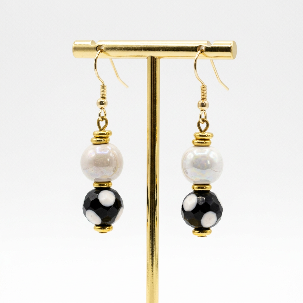 Sefone artisan earrings hanging from a gold-tone display stand, with cream upper beads, black lower beads with white spots, and gold-tone hooks and accents.