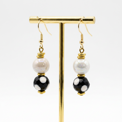 Sefone artisan earrings hanging from a gold-tone display stand, with cream upper beads, black lower beads with white spots, and gold-tone hooks and accents.