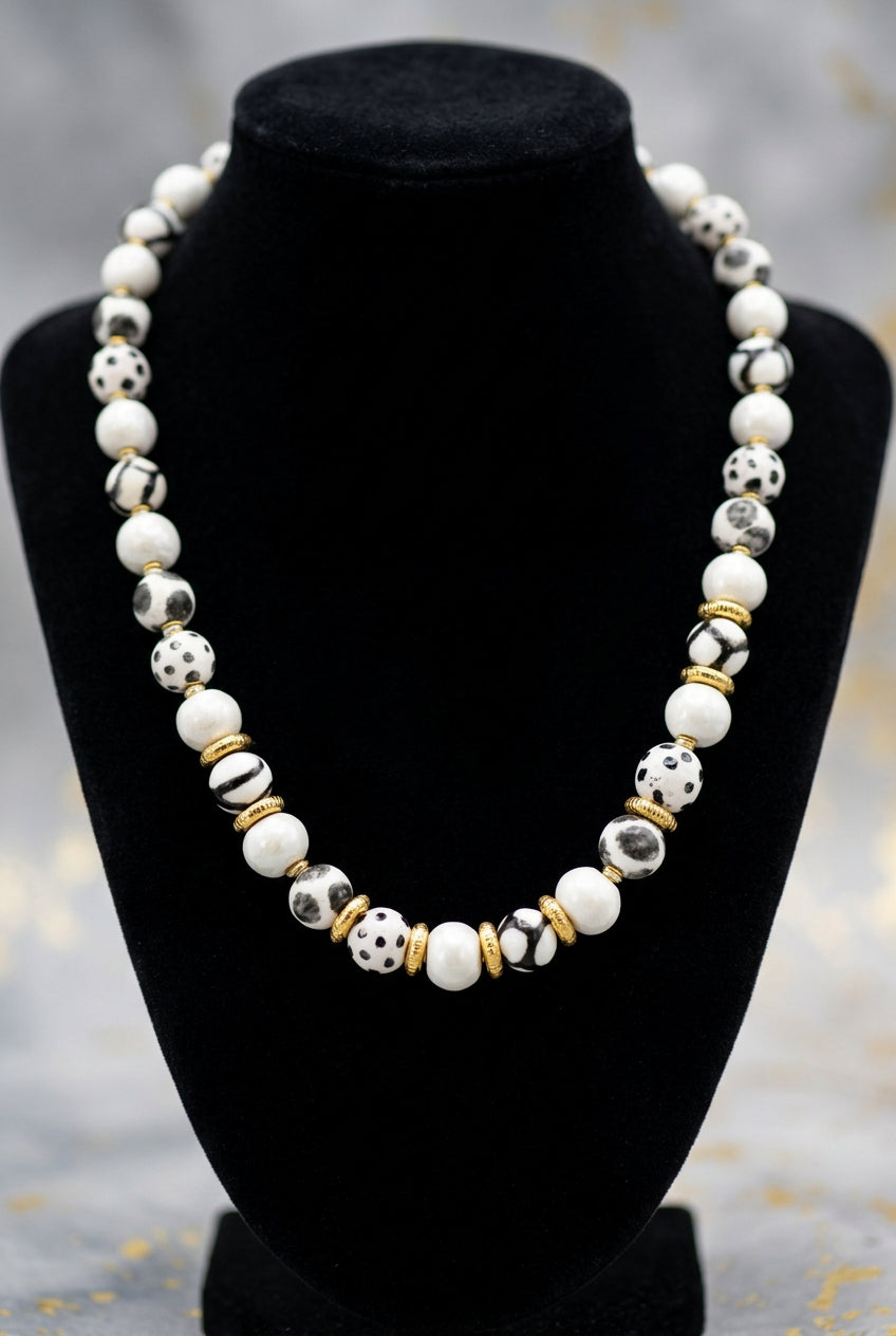 Sefone artisan necklace on a black display bust, showing cream clay beads, spotted and abstract black-and-white patterns, and gold-tone spacers.