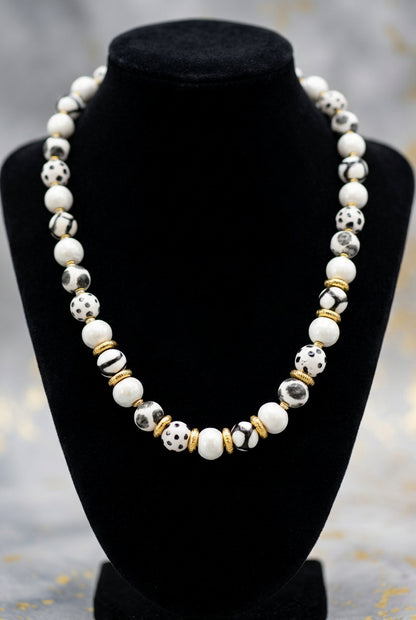 Sefone artisan necklace on a black display bust, showing cream clay beads, spotted and abstract black-and-white patterns, and gold-tone spacers.