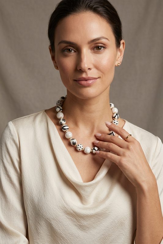 Model wearing a Sefone artisan necklace with cream and patterned black-and-white clay beads over a cream draped-neck blouse, with one hand near the beads.