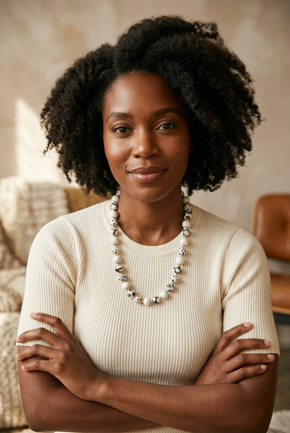 Model wearing a Sefone artisan necklace with cream and patterned black-and-white beads over a cream ribbed short-sleeve top, seated with arms crossed.