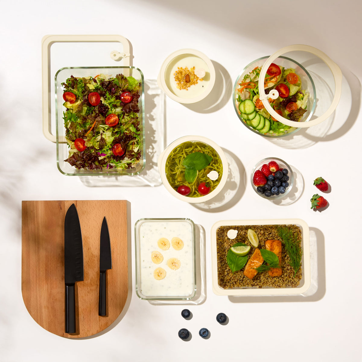 Premium glass food storage set styled in overhead flatlay with fresh salad salmon grain bowls berries and wood cutting board on white surface