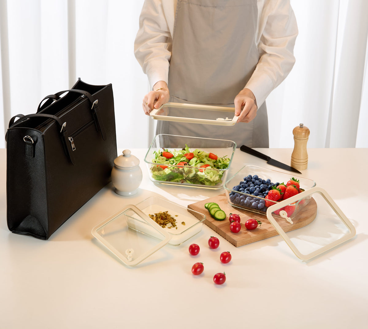Premium glass food storage set in use with person sealing lid over fresh salad container alongside berries tomatoes and black tote bag on white kitchen counter