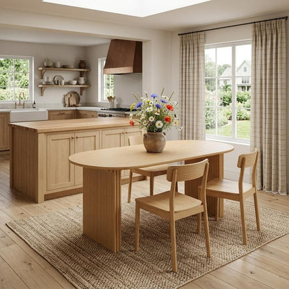 Solid wood double pedestal oval dining table in natural oak finish styled in a farmhouse kitchen dining space with oak chairs, jute rug, and wildflower centerpiece

