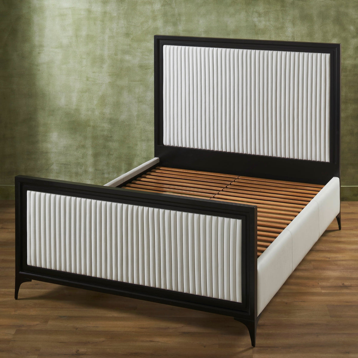 White genuine leather quilted bed frame with matte black border shown without mattress revealing wooden slat support on metal frame, panel headboard and footboard with vertical stripe quilting, iron legs, on hardwood floor against green textured wall. 