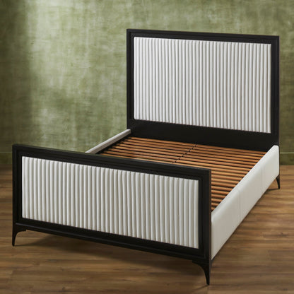 White genuine leather quilted bed frame with matte black border shown without mattress revealing wooden slat support on metal frame, panel headboard and footboard with vertical stripe quilting, iron legs, on hardwood floor against green textured wall. 
