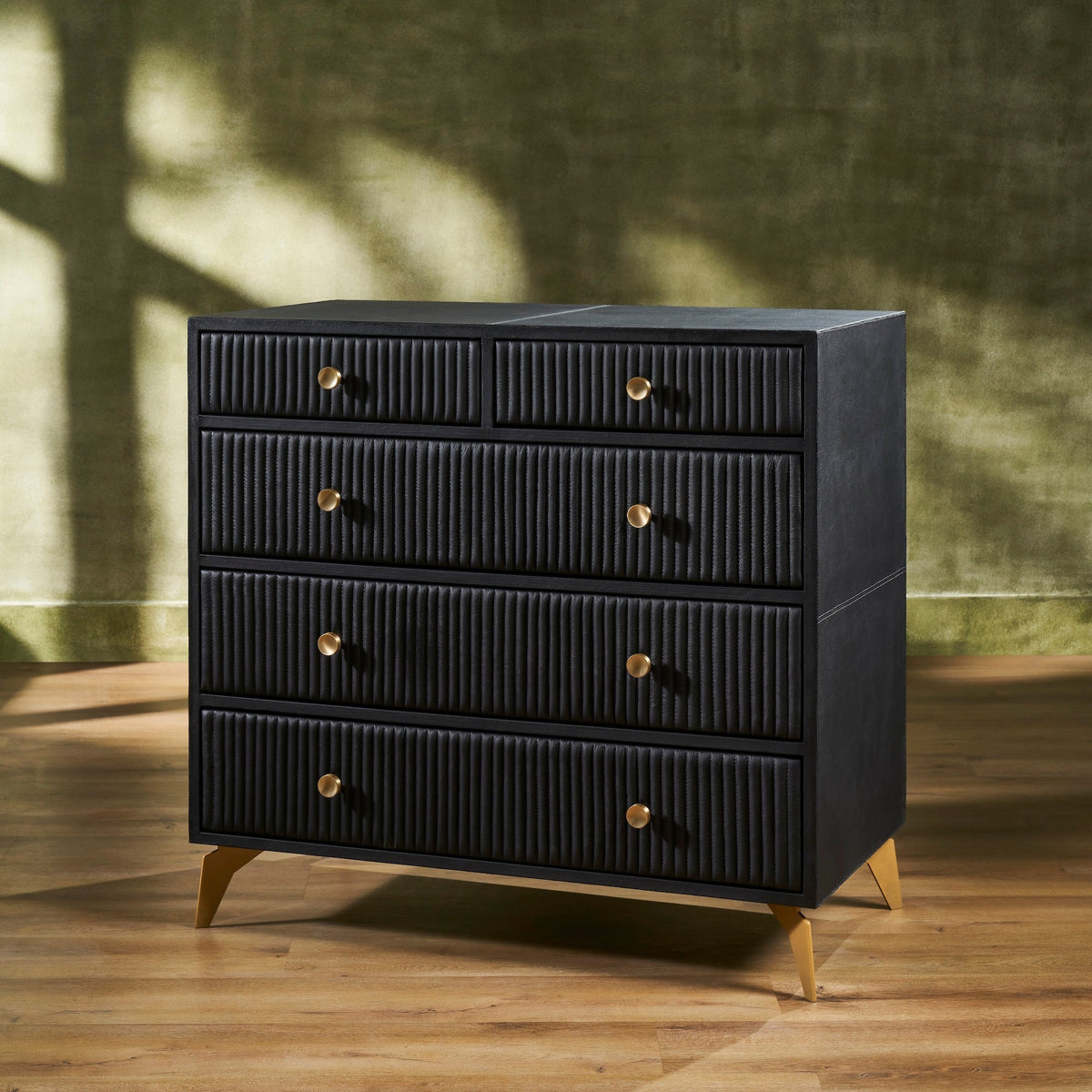 Stanford Ribbed Five-Drawer Leather Chest in black genuine leather at three-quarter angle showing ribbed drawer fronts, brass knobs, and angled brass legs on hardwood floor