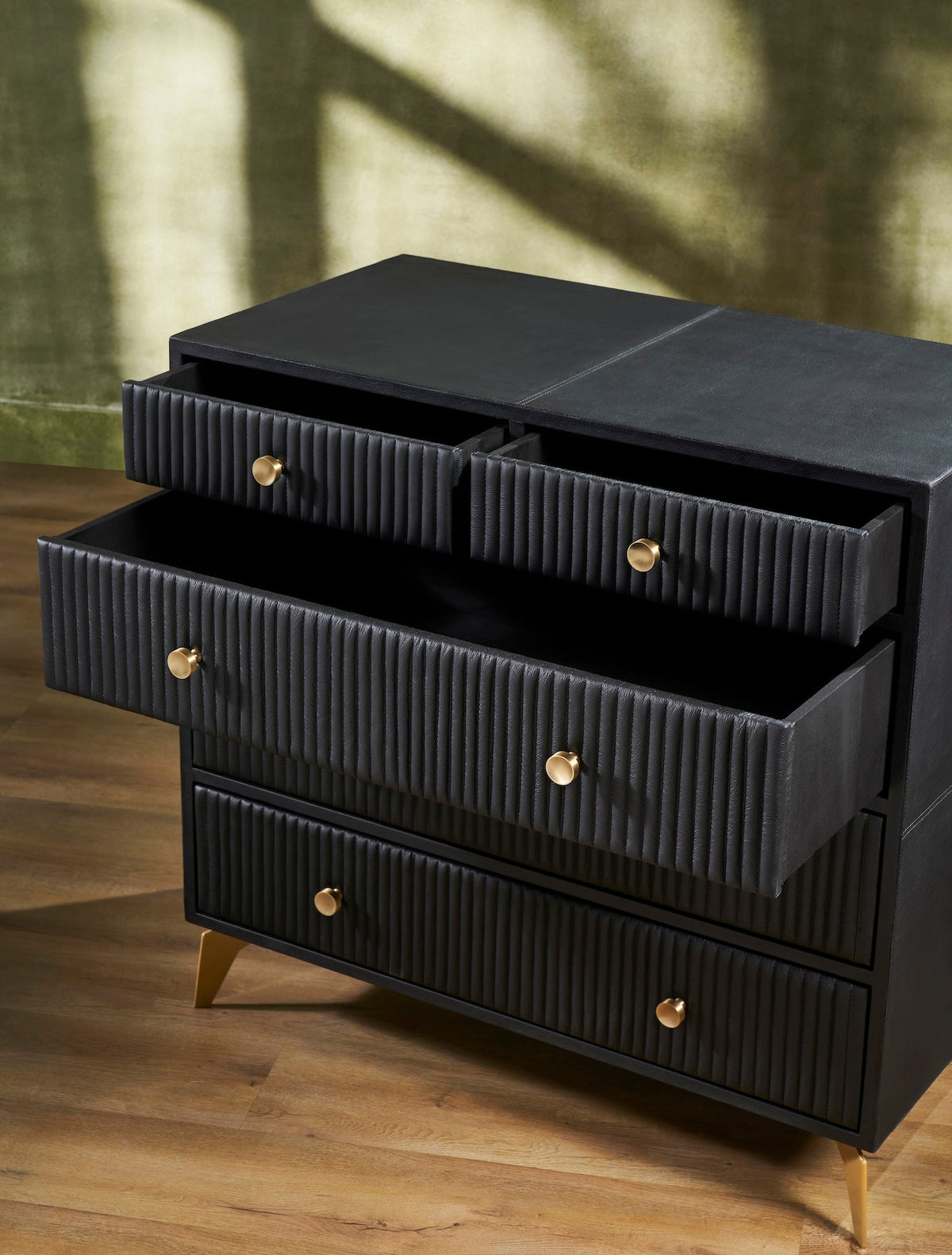 Stanford Ribbed Five-Drawer Leather Chest in black genuine leather with top drawers open showing micro-suede lined interiors and brass knobs on angled brass legs on hardwood floor