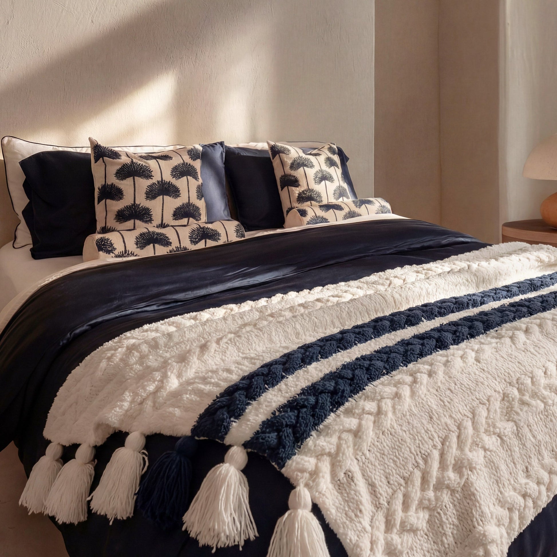Palm leaf embroidered throw pillow navy off white variant styled on made bed with navy velvet duvet white tasseled throw blanket blue satin pillow and warm morning light on textured wall