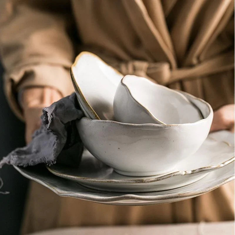 A person holding a stack of the Thetis Tableware Ceramic Set, showcasing the hand-thrown style bowls and stackable ceramic plates with organic edges for a premium hosting feel.