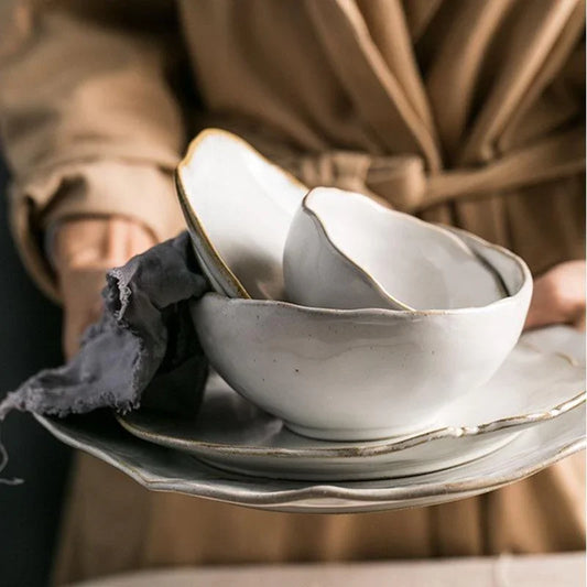 A person holding a stack of the Thetis Tableware Ceramic Set, showcasing the hand-thrown style bowls and stackable ceramic plates with organic edges for a premium hosting feel.