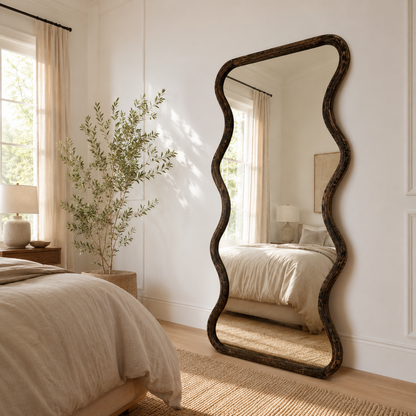 Wavy mango wood wall mirror styled in a bright bedroom beside an olive tree with brown antique frame leaning against white wall

